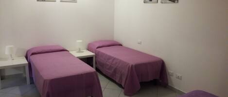 A room with two single beds, each with a purple bedspread, a bedside table with a lamp, and a wall-mounted artwork above the beds.
