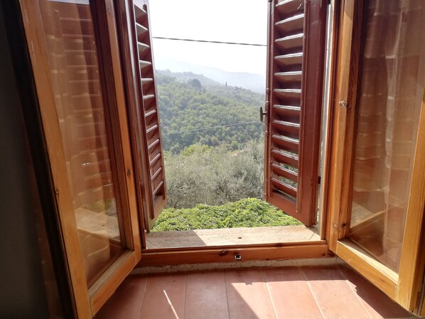 A balcony with wooden shutters open, offering a view of a forested hillside.