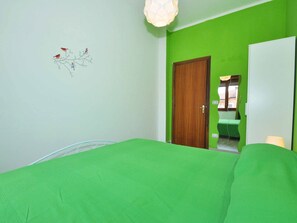 Une pièce avec un mur vert, une porte en bois, un lit blanc recouvert d’une couverture verte, et une décoration murale représentant des oiseaux perchés sur une branche.
