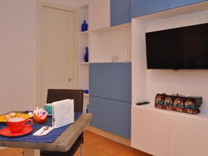 Une pièce avec une table à manger, dressée avec une tasse rouge, une assiette blanche et une cuillère. On y trouve une chaise, une étagère bleue ornée d’objets décoratifs et un téléviseur fixé au mur.