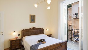 Une chambre avec un lit en bois, une table de chevet, une commode, un lustre, un tableau encadré au mur et un balcon avec une chaise.