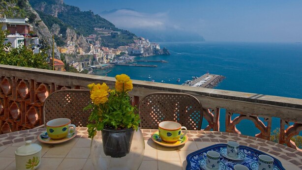 Una mesa con una planta en maceta, dos tazas amarillas y una bandeja con tazas azules y blancas, con vistas a un pueblo costero con edificios y un mar azul cristalino.