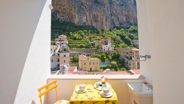 A balcony with a table set for two, overlooking a village with buildings and a cliff. - Appartamento a Amalfi ID 3062 (Amalfi)