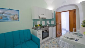 A compact kitchen with white cabinets, a stove, and a sink. There's a dining table set with plates and glasses. A teal sofa is in the living area. - Appartamento a Amalfi ID 3062 (Amalfi)