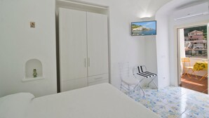 A bedroom with a bed, wardrobe, and a small table with a chair. There's a picture on the wall and a view of buildings through the window. - Appartamento a Amalfi ID 3062 (Amalfi)