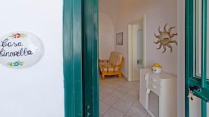 A hallway with a teal door, a sign reading "Casa Kinavella", a wicker chair, and a cabinet with a yellow container.