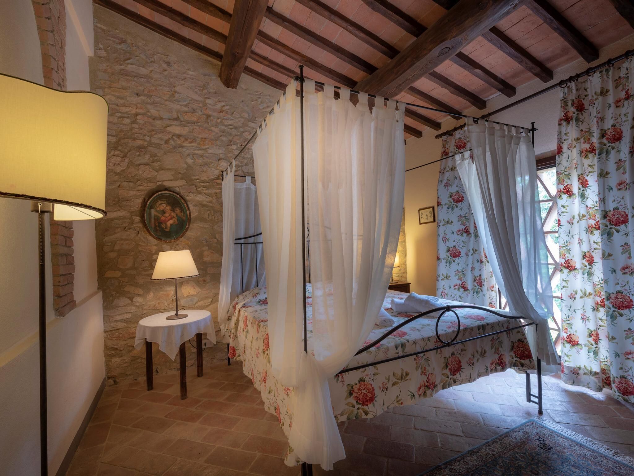 A bedroom with a stone wall, a bed with floral curtains, a bedside table with a lamp, and a rustic wooden ceiling.