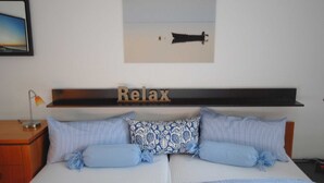 Un dormitorio con cama, almohadas, una mesita de noche, una lámpara, una estantería con un cartel que dice 'Relax' y un cuadro en la pared.