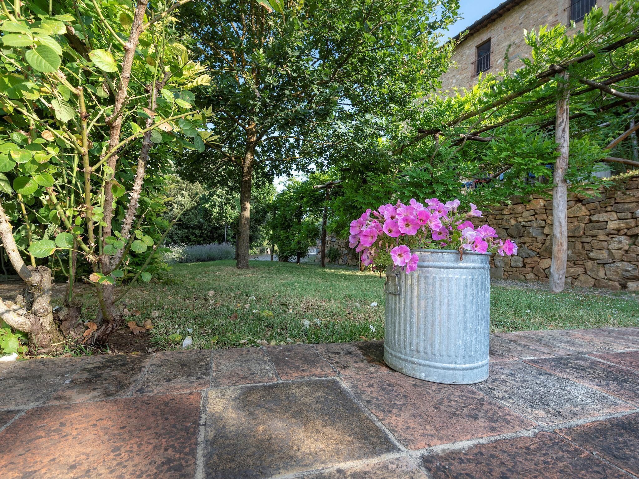A garden with a stone wall, a metal planter with pink flowers, and a building in the background.