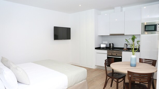 Ett modernt hotellrum med en säng, ett matbord med stolar, ett köksutrymme med vitvaror och en platt-TV som  är monterad på väggen.