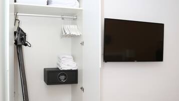 Habitación de hotel con un armario ordenado que contiene toallas dobladas, un secador de pelo y un baúl de almacenamiento. Un televisor de pantalla plana está montado en la pared y se ve una cama con una colcha estampada en verde.
