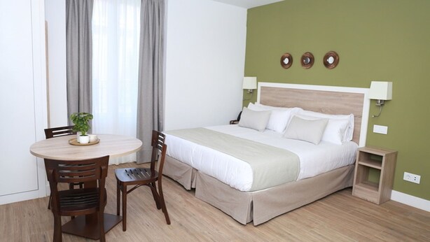 Une chambre d’hôtel avec un lit, une table ronde entourée de chaises, une plante et une petite table d’appoint en bois.