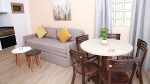 A modern living room with a sofa, two chairs, a small table, and a dining table with chairs. There is a kitchen area with a microwave and oven. A painting is on the wall, and a window with curtains is visible.