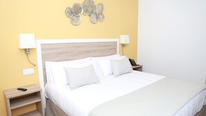A neatly made bed with white linens and a light-colored throw, a wooden nightstand with a remote control, and a wall-mounted decorative piece.