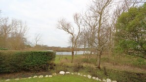 A grassy area with a stone border, trees, and a body of water in the background. - Duck nest at the village pond (Wenningstedt-Braderup (Sylt))