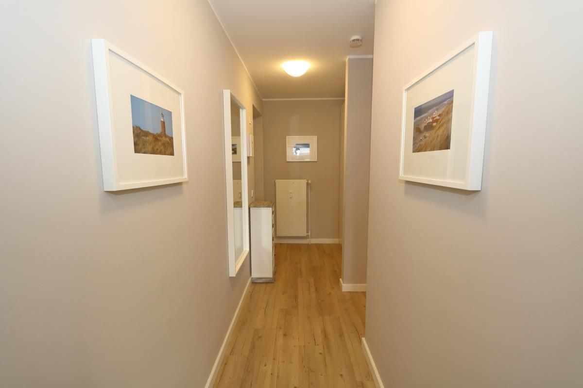 A narrow hallway with wooden flooring and beige walls, flanked by framed pictures on the walls.