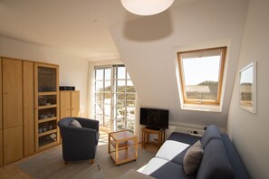 Een woonkamer met een bank, een fauteuil, een televisie, een kast en een groot raam met uitzicht.