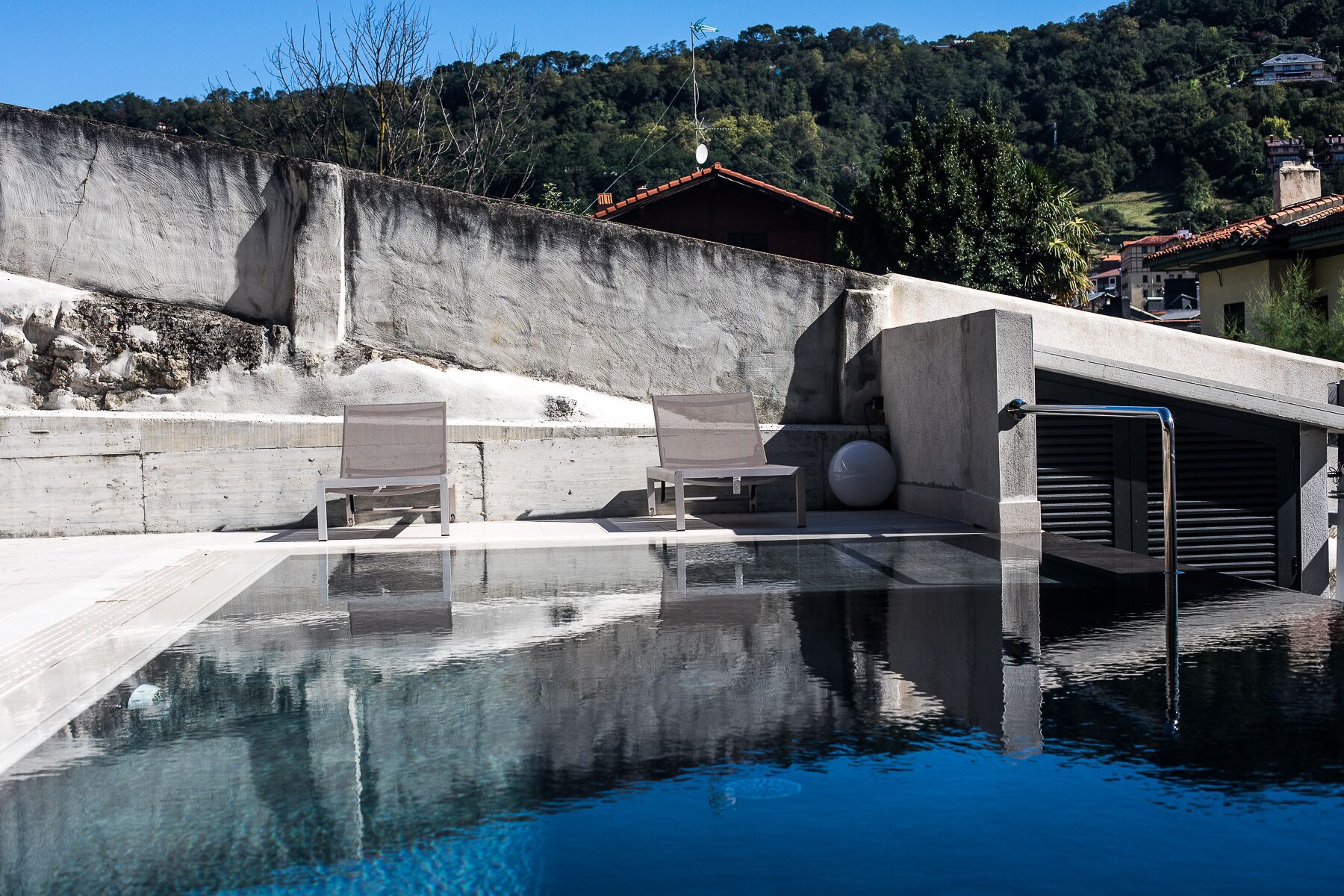 Una piscina con bordo in cemento, una sedia a sdraio e un edificio con tetto rosso sullo sfondo.