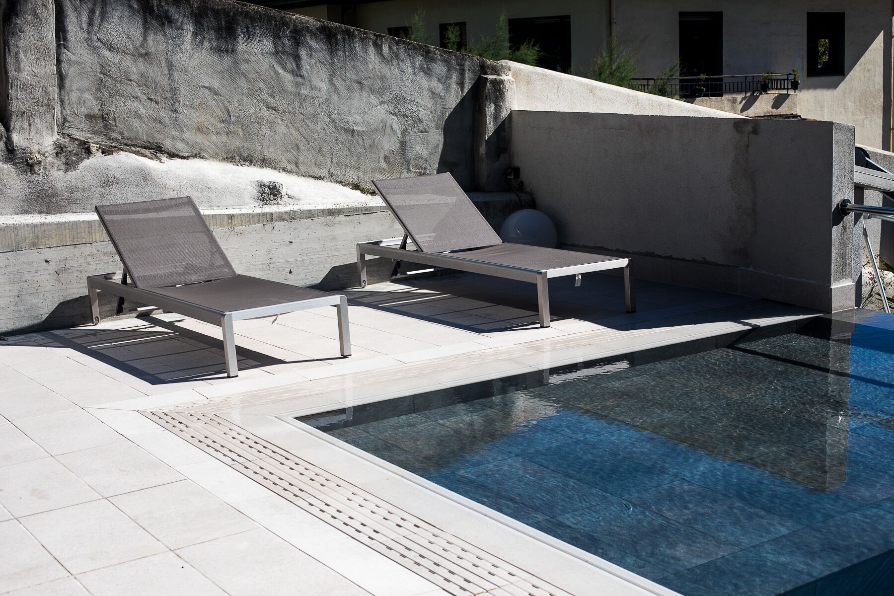 Un área junto a la piscina con dos sillones de descanso y una pequeña mesa, un muro de concreto y una piscina azul cristalina.