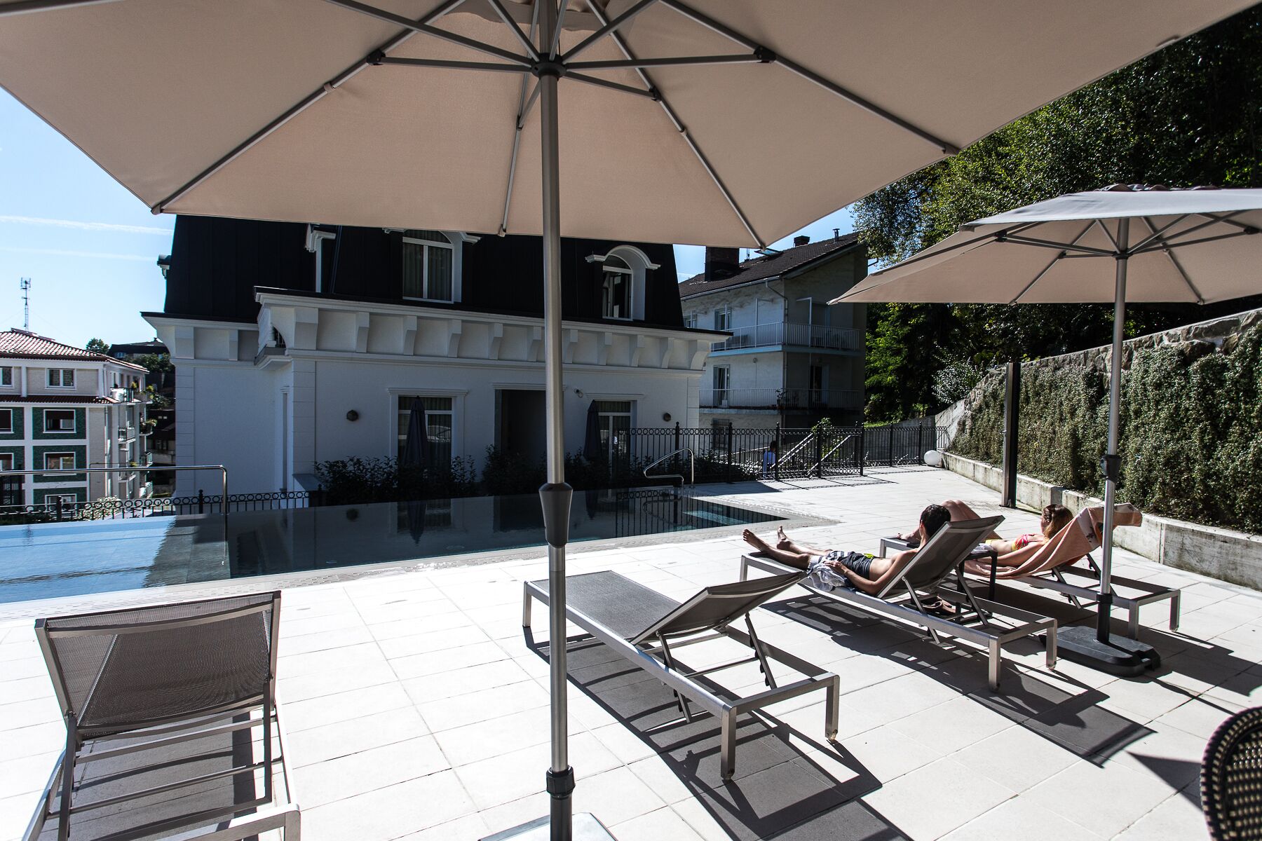 A poolside area with lounge chairs and umbrellas, a swimming pool, and a building with balconies in the background.