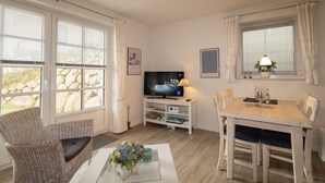 Ett vardagsrum med en tv, ett matbord, flätade stolar, ett soffbord med en vas och stora fönster med gardiner.