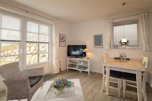 Een woonkamer met een televisie, een eettafel, rieten stoelen, een salontafel met een vaas, en grote ramen met gordijnen.