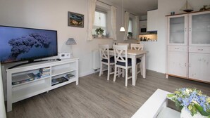Ett modernt vardagsrum med en platt-TV, en matplats med bord och stolar, ett köksområde med skåp och ett vitt konsolbord med böcker och en lampa.