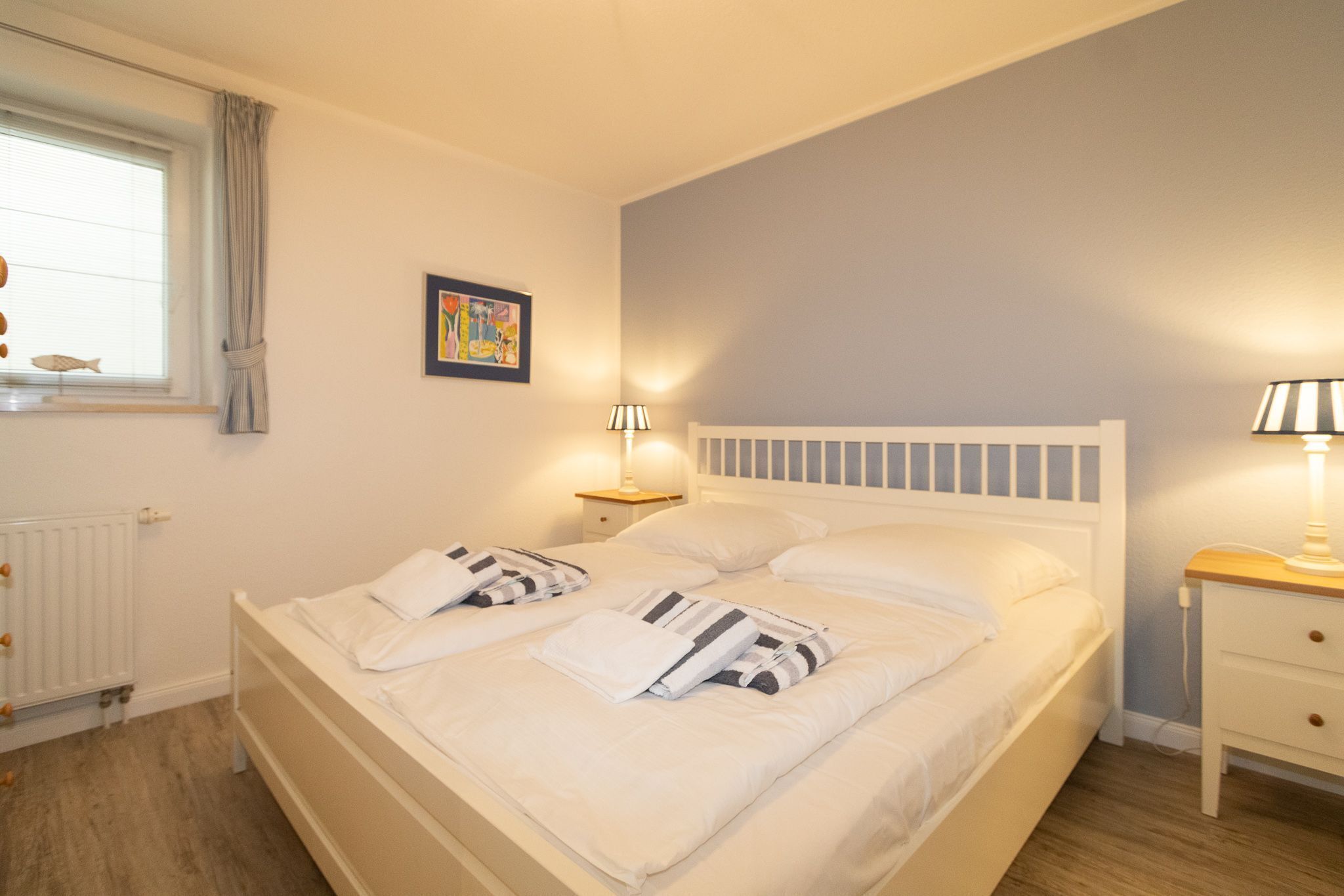 A double bed with white bedding and striped pillows, a bedside table with a lamp, a window with a view of a fish, and a framed picture on the wall.