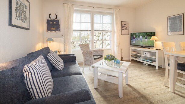 Ett vardagsrum med en grå soffa, ett vitt soffbord, en fåtölj i rotting, en tv och en bokhylla.