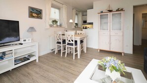 Ett modernt vardagsrum med en platt-TV, en matplats med bord och stolar, ett skåp med glasdörrar och en blomvas på ett soffbord.