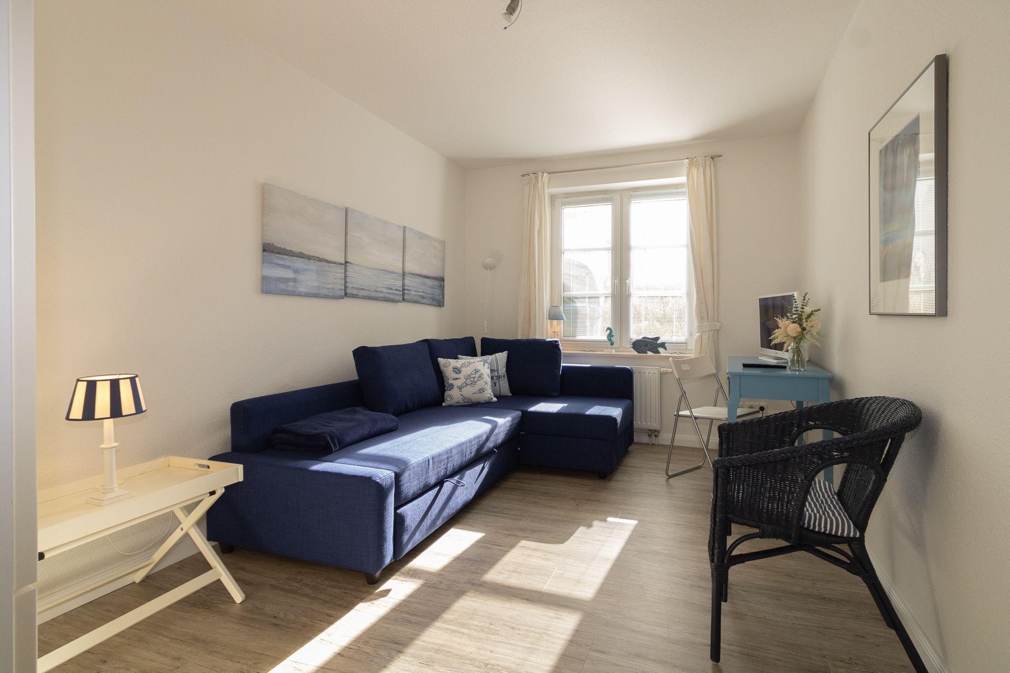 A modern living room with a blue sectional sofa, a small table with a lamp, a wicker chair, and artwork on the walls.