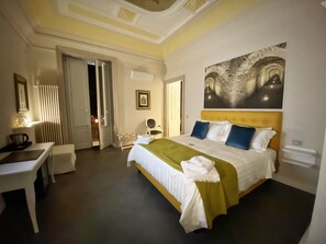 Habitación de hotel con una cama grande, un escritorio y una silla. Hay un cuadro en la pared y una puerta que da a otra habitación.