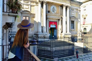 Una mujer está de pie en un balcón con vista a un edificio histórico de arquitectura clásica, que incluye columnas y una fachada detallada.