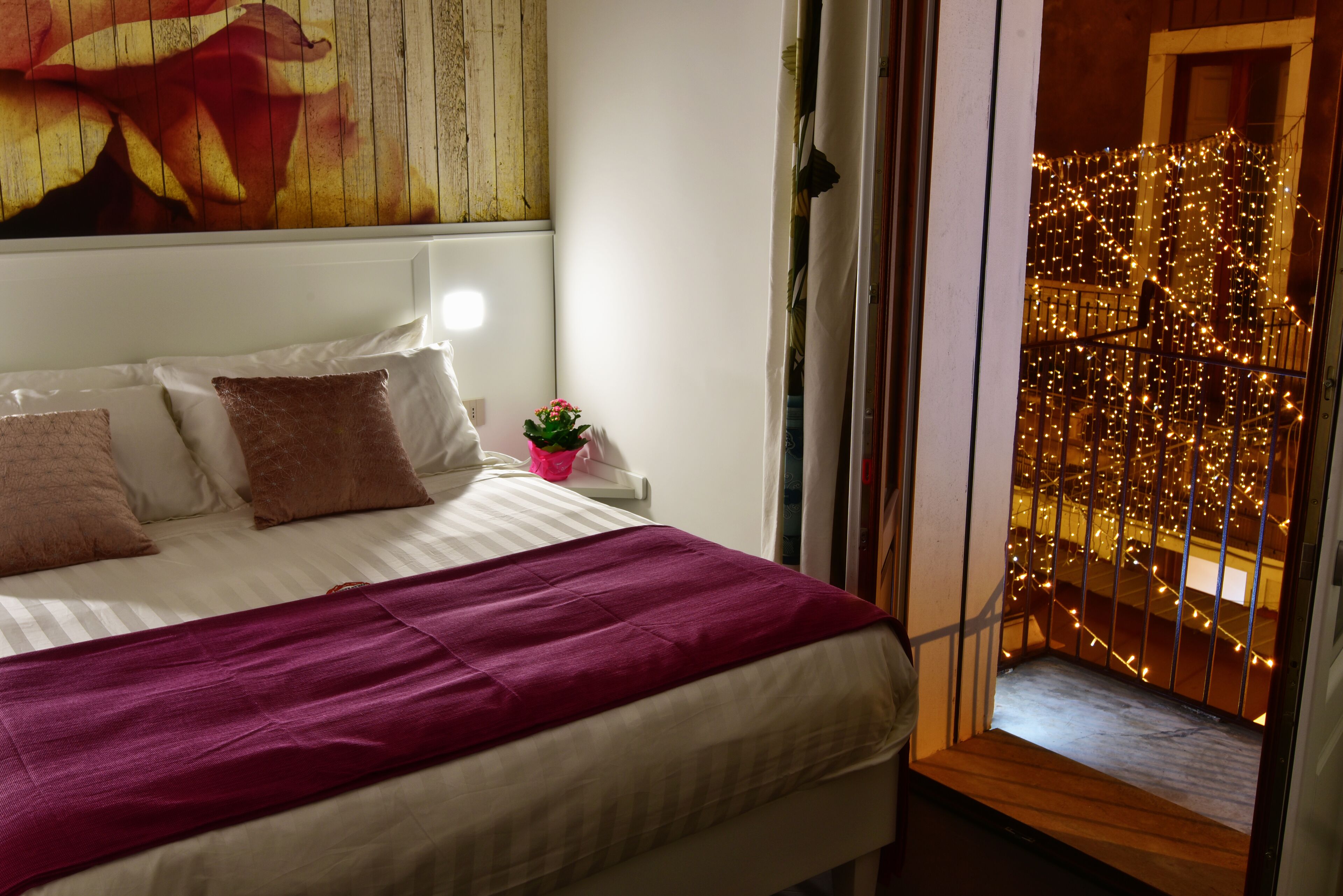 A hotel room with a bed, pillows, a nightstand with a lamp, a painting on the wall, and a view of a balcony with string lights.