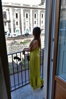 Un balcón con barandilla de metal, una persona con un vestido amarillo y vistas a un edificio con columnas y ventanas.