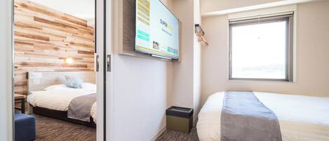 A hotel room with two beds, a wooden headboard, a TV mounted on the wall, a window with blinds, and a carpeted floor.