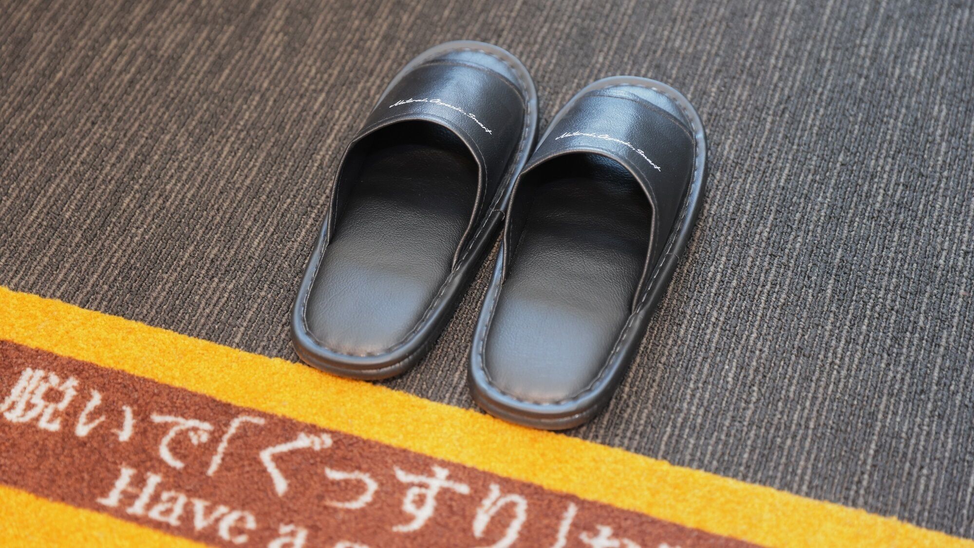 A pair of black shoes on a textured surface with a yellow and brown striped border and Japanese text.
