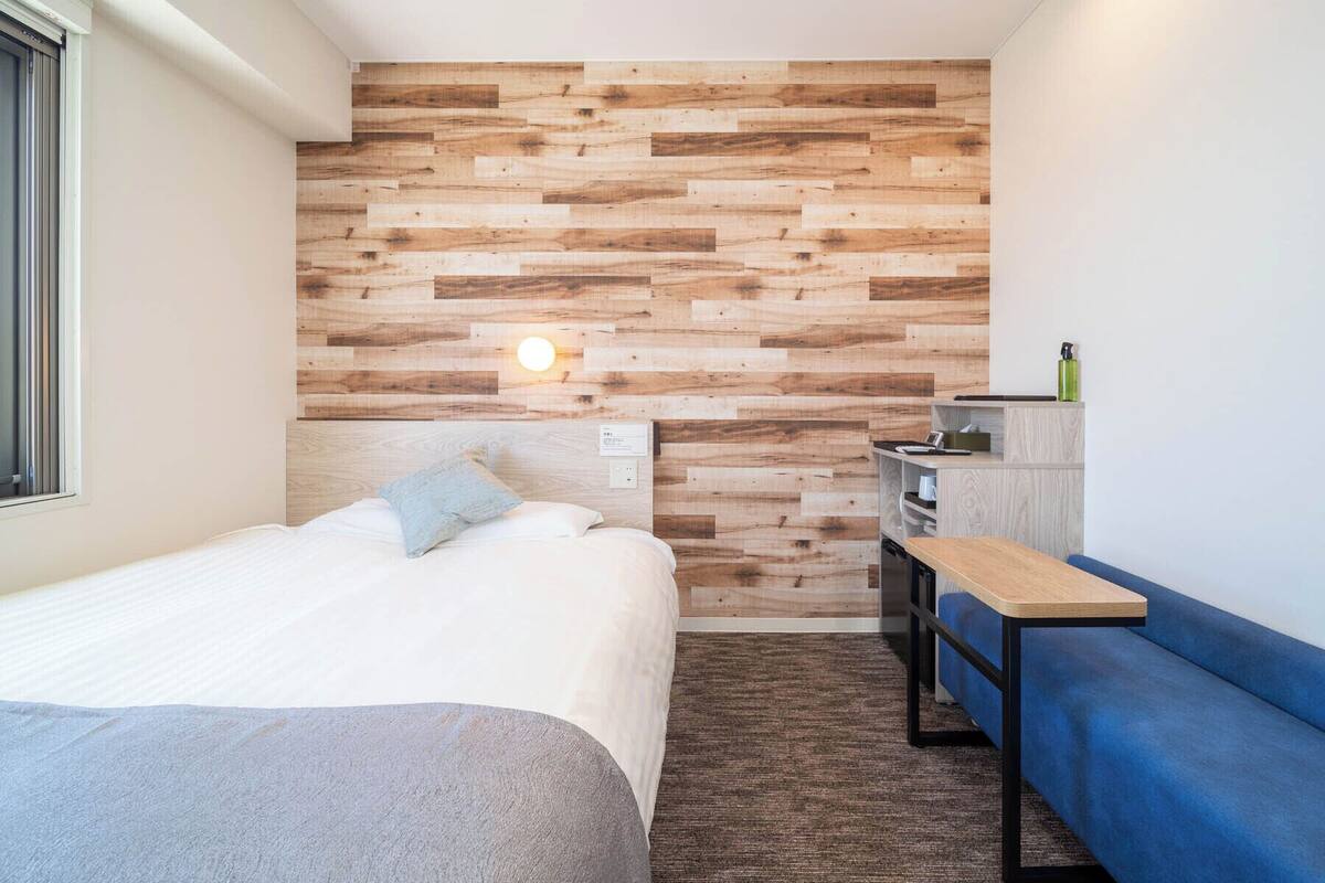 A modern hotel room with a wooden accent wall, a bed with white linens, a blue sofa, a small table, and a nightstand with a lamp.