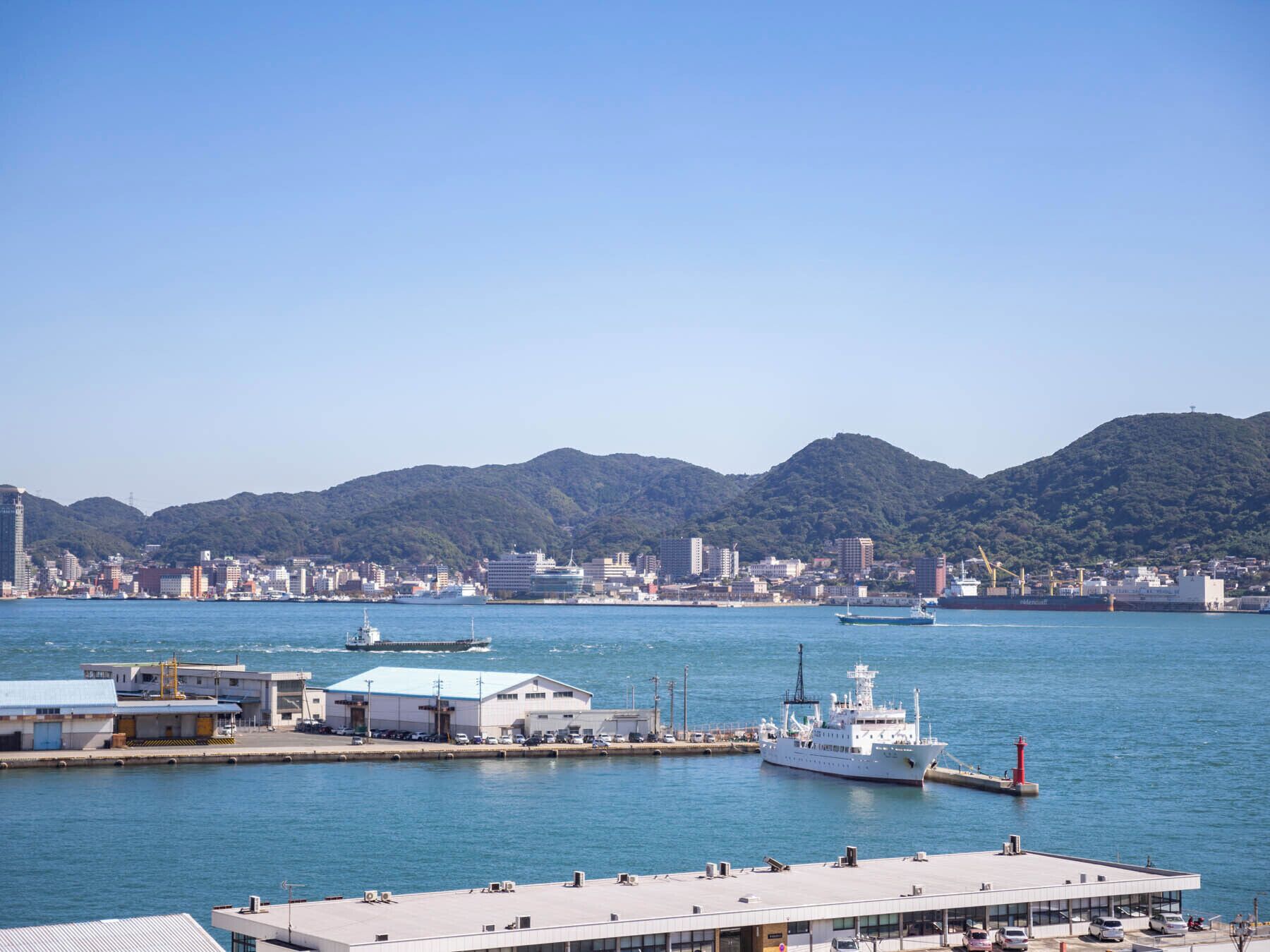 Un puerto con barcos, un muelle y un paisaje urbano con edificios y colinas al fondo.