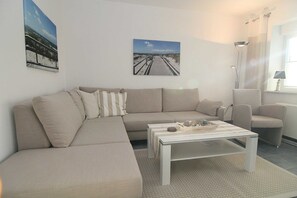 A living room with a beige sectional sofa, a white coffee table, and two armchairs. There are two framed pictures on the wall.