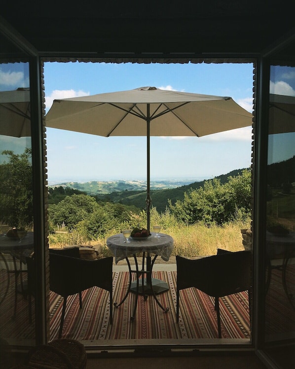 A table with an umbrella, two chairs, and a view of the countryside through a glass door.