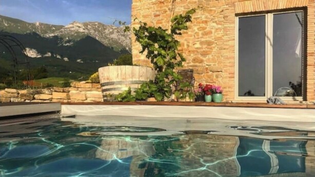 Une piscine à l’eau claire, un bâtiment en pierre doté d’une grande fenêtre et, en arrière-plan, une vue sur la montagne.