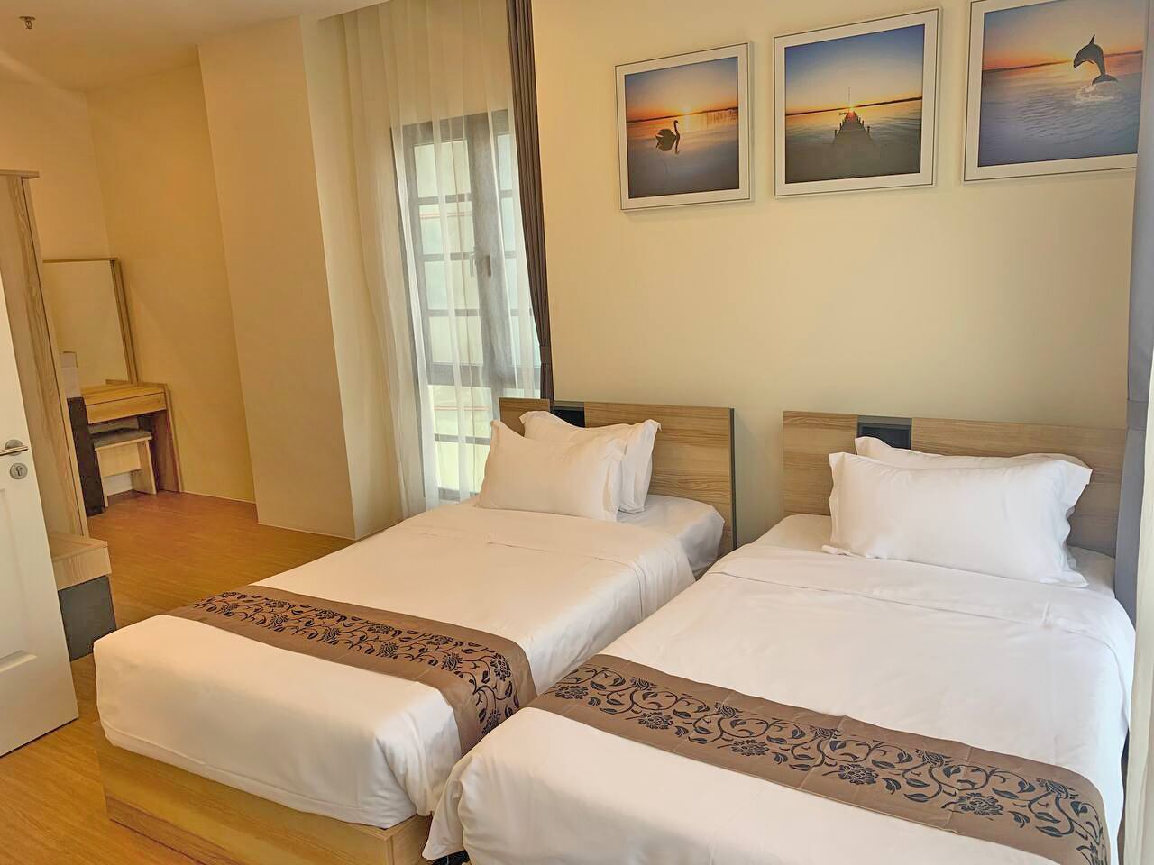 A hotel room with two beds, a wooden floor, a door, a window with curtains, and three framed pictures on the wall.