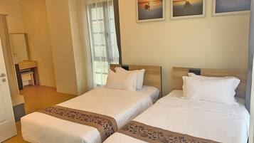 A hotel room with two beds, a wooden floor, a door, a window with curtains, and three framed pictures on the wall.