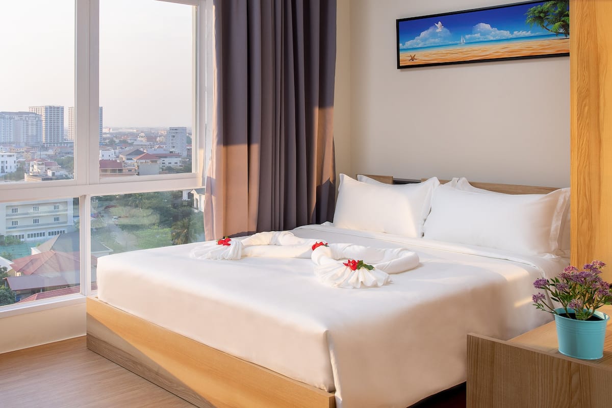 Habitación de hotel con un ventanal grande, una cama con ropa blanca, un cuadro en la pared y una planta pequeña en la mesita de noche.