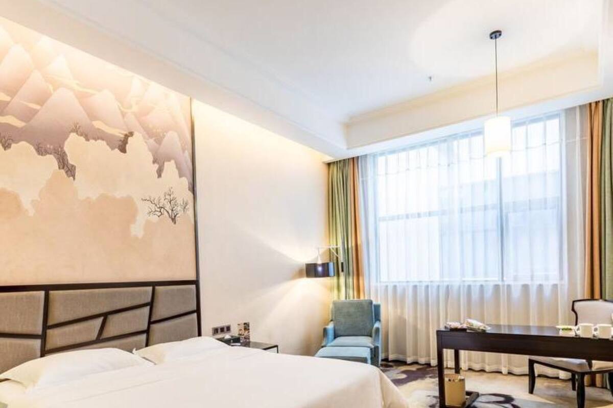 Et hotelværelse med en stor seng, et skrivebord med stol, en lampe og et vindue med gardiner.