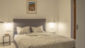 Une chambre avec un grand lit, deux tables de chevet, une commode, un tableau encadré au mur et une porte sur le côté droit.