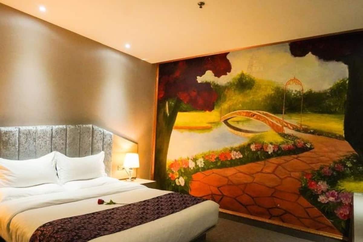 A hotel room with a large bed, a headboard, a bedside table with a lamp, and a mural of a bridge over a river.