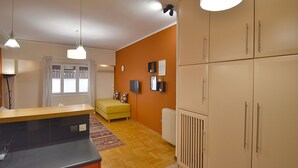 A modern kitchen with a black countertop, wooden flooring, and a yellow sofa. There are cabinets, a radiator, and a window with curtains. - Check Point - Down Town (Athens)