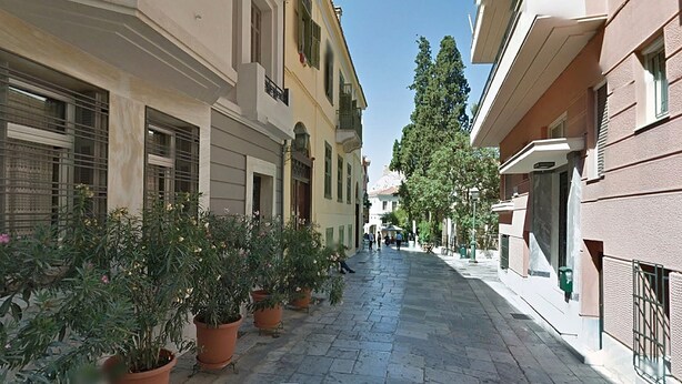A narrow cobblestone street lined with buildings, potted plants, and a few pedestrians. - Check Point - Down Town (Athens)